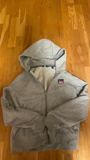 Grå hoodie från GANT med dragkedja - Snygg ljusgrå hoodie från GANT med huva och hel dragkedja framtill. Väldigt skönt material. Tröjan har ett broderat GANT-emblem på bröstet och mjukt foder på insidan. Klassisk passform med långa ärmar och ribbade muddar. Perfekt för chill dagar. 