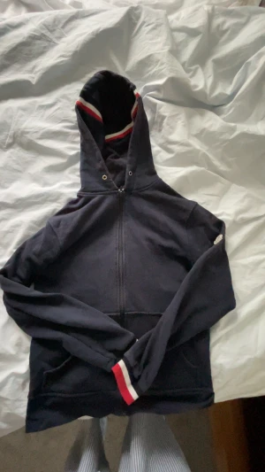 Mörkblå Moncler hoodie XS - Snygg mörkblå hoodie från Moncler i bomull med dragkedja framtill och huva. Röd och vit randig detalj på huva och ärmslut samt klassisk Moncler-logga på ena ärmen. Perfekt för dig som gillar stilrena och sportiga plagg.