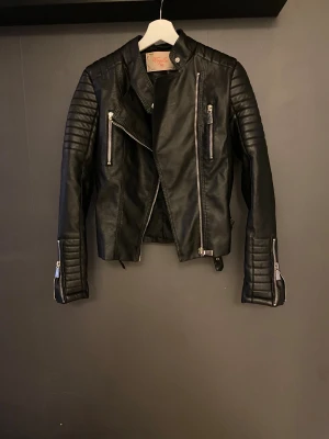 Svart skinnjacka med dragkedjor - Snygg svart skinnjacka från Vogella med coola quiltade detaljer på axlar och ärmar. Jackan har flera silvriga dragkedjor, snedställd front och bältesdetaljer vid midjan. Perfekt för dig som gillar en edgy stil och vill sticka ut.