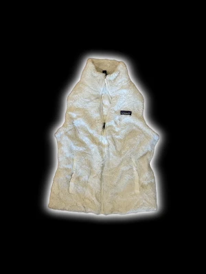 Vit pälsväst från Patagonia - Säljer en fluffig vit pälsväst från Patagonia med hög krage och dragkedja framtill. Västen har två fickor med dragkedja och är fodrad med slätt tyg på insidan. Perfekt för lager-på-lager och har en clean, modern look.