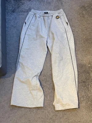 Grå mjukisbyxor Broken Planet XL - Grå mjukisbyxor från Broken Planet i storlek XL. Byxorna har bred passform, elastisk midja och dragkedjor på fickorna. Snyggt kamouflagemönstrat BP-logga på höften. Perfekta för chill eller streetwear.