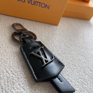 Vuitton svart väskdekoration - Snygg svart väskdekoration från  Vuitton i skinn med metallfäste och ikoniska loggan i silver. Perfekt att hänga på din väska för att ge den en lyxig och personlig touch. Stilren och modern design med exklusiva detaljer.