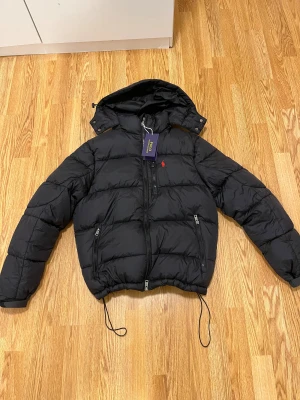 Svart pufferjacka från Polo Ralph Lauren - Svart pufferjacka från Polo Ralph Lauren med huva, dragkedjor och snörning nedtill. Jackan har det klassiska röda Polo-logot på bröstet och två sidofickor med dragkedja. Materialet är syntet och jackan är perfekt för kalla dagar.