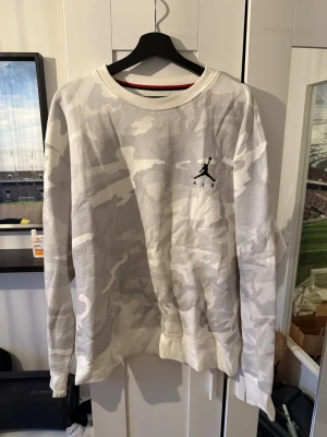 Jordan Air camo sweatshirt i grått - Säljer en oversized crewneck från Jordan Air med grått och vitt camo-mönster. Tröjan har rund hals, lång ärm och broderad Jumpman-logga på bröstet. Perfekt för dig som gillar streetwear och vill sticka ut med en clean look.