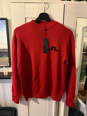 Röd stickad tröja från J.Lindeberg - Röd stickad tröja från J.Lindeberg med svart logga på bröstet. Tröjan har ribbad krage, ärmslut och nederkant samt lång ärm. Perfekt för dig som vill ha en clean och stilren look med en touch av sportig vibe.