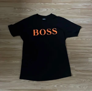Svart BOSS t-shirt med orange tryck - Svart t-shirt från Hugo Boss med stort orange BOSS-tryck på bröstet och diskret HUGO BOSS-text under. Klassisk rund hals och korta ärmar. Tillverkad i 100% bomull för skön och mjuk känsla. Perfekt för dig som gillar stilrena och tydliga loggor.