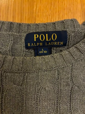 Grå stickad tröja från Polo Ralph Lauren - Snygg grå kabelstickad tröja från Polo Ralph Lauren med klassisk rund hals och långärmad design. Tröjan har det ikoniska broderade Polo-logot på bröstet och ribbade muddar vid ärmslut och nederkant. Perfekt för dig som gillar stilren och tidlös look.