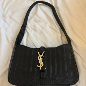 Svart axelväska från Yves Saint Laurent - Snygg svart axelväska från Yves Saint Laurent med krokodilpräglat skinn och guldigt YSL-emblem framtill. Väskan har justerbar axelrem med gulddetalj och dragkedja upptill. Perfekt storlek för det viktigaste och ger en lyxig vibe till din outfit. Villig att gå ner i pris vid snabb affär❤️