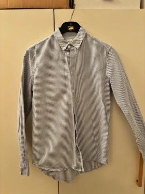 Randig skjorta från Samsøe Samsøe - Klassisk skjorta från Samsøe Samsøe i vitt och blått med smala ränder. Skjortan har lång ärm, knappar framtill och krage. Tillverkad i mjuk bomull, perfekt för en clean och stilren look.