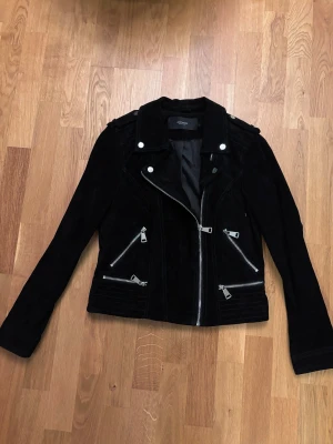 Svart mockajacka från Vero Moda - Snygg svart mockajacka från Vero Moda med coola silverfärgade dragkedjor och detaljer. Jackan har lång ärm, klassisk krage och flera fickor med dragkedja framtill. Perfekt för dig som vill ha en edgy look och gillar stilrena detaljer.