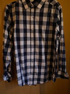 Blårutig skjorta med knappar - Snygg skjorta med stort blårutigt mönster och klassisk krage. Skjortan har långa ärmar med manschett och vita knappar framtill. Perfekt för dig som gillar en stilren och modern look. Materialet känns lätt och bekvämt mot huden.