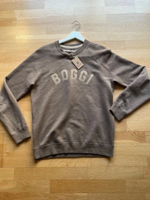 Beige cashmir tröja från Boggi Milano  - Snygg beige sweatshirt från Boggi Milano i Kashmirblandning med broderad logga framtill. Tröjan har rund hals, ribbade muddar vid ärmslut och nederkant samt långärmad design. Perfekt för en chill och stilren look. Nypris: 1900