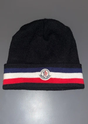 Svart Moncler mössa - Svart mössa från Moncler med bred uppvikt kant i blått, vitt och rött. Klassisk Moncler-logga framtill. Stickad i mjukt material som känns skönt mot huvudet. Perfekt för dig som vill ha en clean look med lite färg.