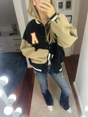 Svart och beige bomberjacka med A-patch - Snygg bomberjacka i svart med kontrasterande beige ärmar och vit-svarta ribbade muddar. Jackan har en stor röd A-patch på bröstet och knäppning framtill. Oversized passform och sportig collegekänsla, perfekt för en trendig streetstyle.
