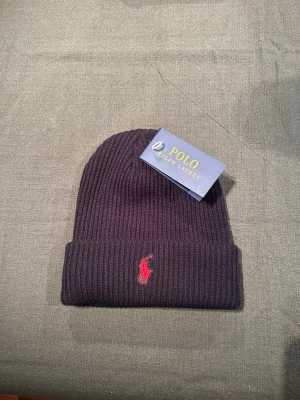 Svart ribbad mössa från Polo Ralph Lauren - Svart ribbstickad mössa från Polo Ralph Lauren med klassisk röd broderad logga framtill. Snygg och enkel design som passar till allt, tillverkad i mjukt material för extra komfort. Perfekt för kalla dagar och en stilren look. Ser lite lila ut på bilden fast den är svart
