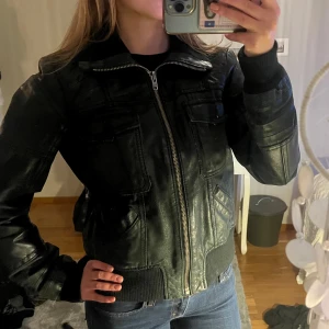 Svart skinnjacka från ONLY, storlek L - Säljer en svart skinnjacka från ONLY i storlek L. Jackan har klassisk bombermodell med dragkedja framtill, flera fickor och ribbade muddar vid ärmslut och midja. Perfekt för dig som vill ha en edgy och tidlös look.