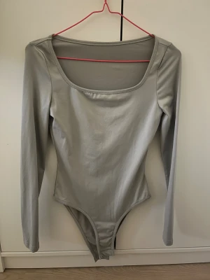 Grå långärmad bodysuit - Snygg grå bodysuit med lång ärm och rundad halsringning. Modellen är tight och har knäppning i grenen för enkel användning. Materialet är mjukt och stretchigt, perfekt för att skapa en clean och stilren look.