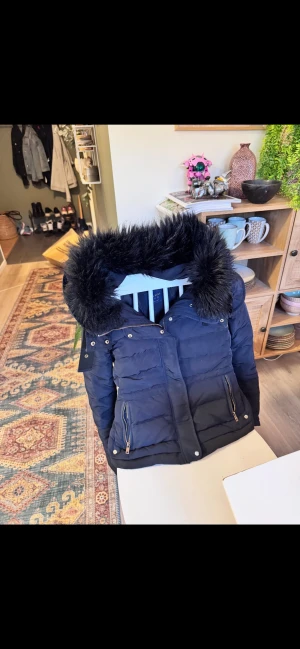 Svart pufferjacka med fuskpäls - Snygg svart pufferjacka med fluffig fuskpäls på huvan och guldfärgade dragkedjor. Jackan har två framfickor med dragkedja och quiltad design som ger en cool look. Perfekt för kalla dagar när du vill vara både varm och stilren. Storlek M men passar för dom med S för jag själv brukar ha S i jackor.