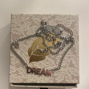 Silverfärgat halsband med DREAM-text - Snyggt halsband i silverfärgad metall med ett hänge där det står DREAM i rosa och silver. Kedjan är klassisk och har ett smidigt lås. Perfekt accessoar för att ge din outfit lite extra attityd.