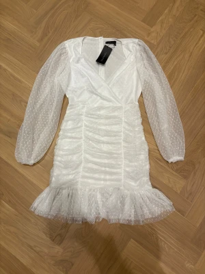 Vit prickig kort klänning Trendyol - Supersöt vit kort klänning från Trendyol med transparenta långärmar och små prickar i tyget. Klänningen har omlott-v-ringning, rynkade detaljer längs kroppen och en volangkant nedtill. Materialet är lätt och meshigt, perfekt för att sticka ut med en romantisk vibe.