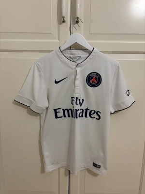 PSG Ibrahimović 10 vit fotbollströja Nike - Säljer en vit Paris Saint-Germain fotbollströja från Nike med Dri-Fit material. Tröjan har korta ärmar, mörkblå detaljer och PSG-logga på bröstet. På ryggen står Ibrahimović och nummer 10 i blått och guld. Fly Emirates tryck på framsidan.