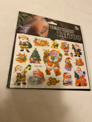 Juliga temporära tatueringar - Säljer ett ark med färgglada temporära tatueringar med julmotiv som tomtar, julgran, presenter, björn och polkagris. Perfekt för att piffa upp din look med roliga och festliga motiv. Tatueringarna är i plastmaterial och appliceras enkelt på huden med vatten.