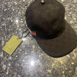 Svart Gucci keps med GG-mönster - Säljer en svart keps från Gucci med klassiskt GG-monogram över hela kepsen. Kepsen har en böjd skärm, justerbar rem bak i skinn och en snygg detalj med grönt och rött band på sidan. Materialet är bomull och skinn, perfekt för dig som gillar lyxiga accessoarer. Pris kan diskuteras.