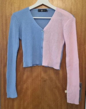 Tvåfärgad ribbad kofta Daisy Street - Säljer en croppad kofta från Daisy Street i ribbad bomullsmix. Ena sidan är ljusblå och andra sidan ljusrosa, med v-ringning och knappar framtill. Långärmad och perfekt för lager-på-lager-stil.