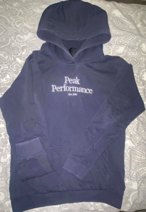 Mörkblå hoodie från Peak Performance - Mörkblå hoodie från Peak Performance med vit broderad logga på bröstet. Klassisk modell med huva använd ett par tillfällen annars sitter jätteskönt snygg att stylea med 