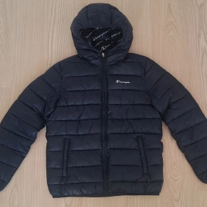 Mörkblå pufferjacka från Champion - Mörkblå vinterjacka från Champion. Bokstaven n saknas på bröstet. Annars väldigt fint skick. Stl 162, 13-14 år. Snygg mörkblå pufferjacka från Champion med huva och dragkedja framtill. Jackan har quiltad design, två sidofickor och logotyp på bröstet samt insidan. Perfekt för kalla dagar och riktigt chill streetstyle.