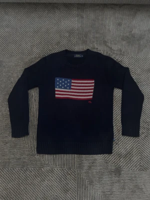 Polo Ralph Lauren knit amerikans flagga - Mörkblå stickad tröja från Polo Ralph Lauren med amerikansk flagga broderad på bröstet och RL-detalj. Klassisk rund halsringning och ribbade muddar vid ärmslut och nederkant. Perfekt för dig som gillar preppy stil.