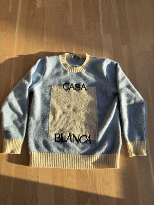 Casa Blanca ljusblå stickad tröja - Snygg ljusblå stickad tröja från Casa Blanca med kontrasterande krage, muddar och en stor beige ruta framtill. Svart broderad text 'CASA BLANCA' på framsidan. Perfekt för dig som gillar unik design och mjuka material.
