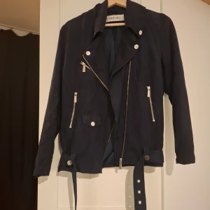 Mörkblå bikerjacka från Ivyrevel - Snygg mörkblå bikerjacka från Ivyrevel med flera silvriga dragkedjor, knappar och bälte i midjan. Jackan har lång ärm och cool asymmetrisk krage. Perfekt för dig som vill ha en edgy stil och gillar detaljer.