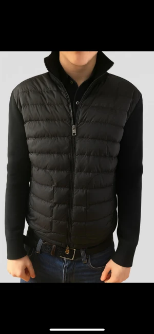 Svart dunväst från Moncler - Snygg svart dunväst från Moncler med quiltad framsida och hög krage. Västen har dragkedja framtill och två dragkedjefickor. Materialmix med dun och stickade partier på sidor och rygg ger en modern look. Perfekt för lager-på-lager-stil.