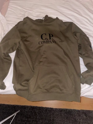 Olivgrön hoodie från C.P. Company - Olivgrön hoodie från C.P. Company med svart logga på bröstet och den klassiska linsen på ärmen. Tröjan har känguruficka framtill och ribbade muddar. Tillverkad i mjuk bomull, perfekt för en chill och stilren look. Perfekt för en skön hoodie nu i vinter och till våren. Tvivla inte att fråga vid funderingar. Priset är inte hugget i sten