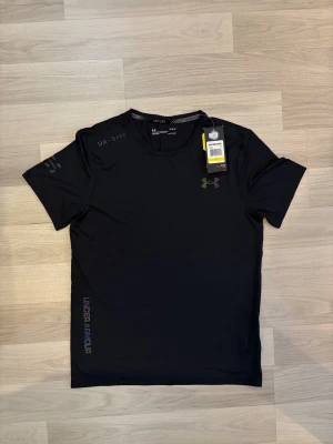 Svart Under Armour HeatGear t-shirt - Svart t-shirt från Under Armour med diskret logga på bröstet och ärmen. Tillverkad i lätt och ventilerande HeatGear-polyester, perfekt för löpning och träning. Kortärmad modell med rund hals och slim passform. Snygga detaljer med UA-SPEC-tryck och färgskiftande text längs sidan.