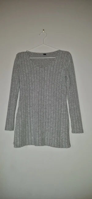 Ribbad stickad tröja - Mysig grå mjuk stickad tröja med ribbat mönster och lång ärm. Tröjan har en lätt v-ringad hals och är lite längre i modellen, perfekt för lager-på-lager. Enkel och stilren design som passar till det mesta.