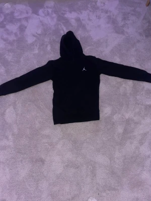Svart hoodie från Jordan med huva - Svart hoodie från Jordan med klassisk Jumpman-logga på bröstet. Tröjan har huva och långa ärmar, tillverkad i mjuk bomull och polyesterblandning. Perfekt för dig som gillar streetwear och sportig stil.