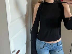 Svart långärmad topp med snörning - Säljer en svart långärmad topp med coola cut-outs på axlarna och snörningsdetaljer längs ärmarna. Toppen är croppad och har en rund halsringning. Materialet känns mjukt och lätt, perfekt för dig som gillar edgy och trendiga plagg.