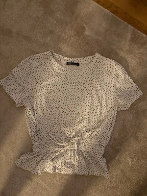 Prickig vit blus från Zara med knyt - Säljer en söt vit blus från Zara med svarta prickar och korta ärmar. Toppen har ett knyt i midjan och en lätt volang nedtill som ger en chill och feminin vibe. Perfekt för dig som gillar prickigt och vill ha något som sticker ut lite extra.
