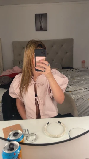 Ljusrosa blus med puffärmar och knyt - Supersöt ljusrosa blus med stora puffärmar och två knyt framtill. Blusen är croppad och har en luftig passform som ger en trendig vibe. Perfekt för dig som gillar statement-ärmar och unika detaljer.