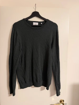 Tröja Bläck - En stilren mörkgrön stickad tröja från Bläck i storlek M. Tröjan har rund halsringning, ribbade muddar och lång ärm. Perfekt för lager-på-lager och enkel att matcha med jeans eller chinos.