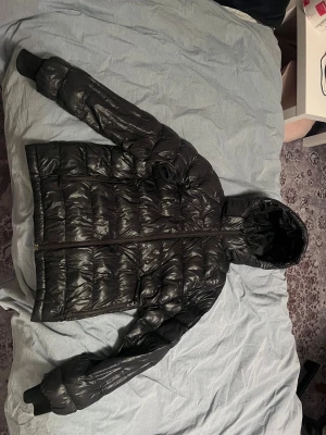 Svart dunjacka Peak Performance 160 - Svart dunjacka från Peak Performance i storlek 160. Jackan har en glansig finish, huva och dragkedja framtill.