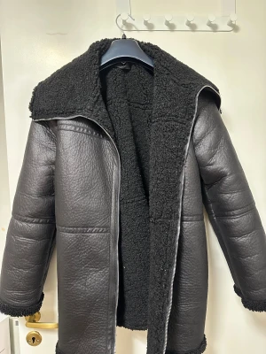 Svart teddyjacka med skinnlook - Säljer en svart teddyjacka med skinnlook och fluffigt foder. Jackan har en rejäl krage, grov dragkedja och detaljer i teddy vid ärmslut och nederkant. Perfekt för dig som vill ha en cool och varm jacka med oversized känsla. 