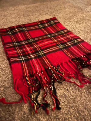 Röd rutig halsduk från Barbour - Säljer en klassisk röd rutig halsduk från Barbour med fransar i ändarna. Halsduken har inslag av svart, vit och gul i mönstret och är tillverkad i mjuk ull. Perfekt för att ge din outfit en färgklick och hålla dig varm under kyliga dagar.