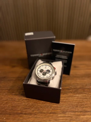 Tommy Hilfiger kronograf herrklocka - Stilren kronografklocka från Tommy Hilfiger med vit urtavla, svarta och röda detaljer samt tre subdials. Boetten och länken är i silverfärgat metall, vilket ger en klassisk och modern vibe. Perfekt accessoar för dig som gillar snygga och funktionella klockor.