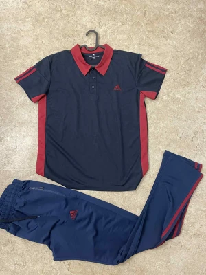 Adidas marinblå träningsset med röda detaljer - Snyggt träningsset från Adidas med marinblå pikétröja och matchande byxor. Tröjan har röd krage, röda ränder på axlarna och sidorna samt Adidas-logga på bröstet. Byxorna har röda ränder längs benen, dragkedja vid fickan och elastisk midja. Perfekt för sportiga dagar.