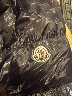 Svart pufferjacka från Moncler XXL - Säljer en mörk blå  pufferjacka från Moncler i storlek XXL. Jackan har glansigt yttermaterial, klassisk Moncler-logga på ärmen och coolt serietidningsfoder på insidan. Dragkedja och tryckknappar framtill. Perfekt för dig som vill ha nått billigt!