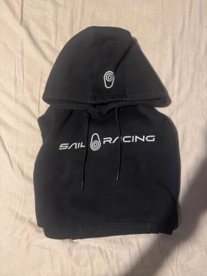 Svart Sail Racing hoodie - Svart hoodie från Sail Racing med stort vitt logotryck i mitten. Perfekt för dig som gillar stilren streetwear och vill ha något bekvämt och snyggt. Dm för mer detaljer