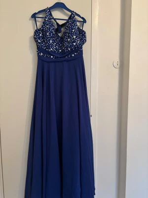 Blå långklänning med paljetter - Säljer en blå långklänning med glittrande paljetter och pärlor på överdelen. Klänningen har halterneck och ett snyggt kors i ryggen. Kjolen är svepande och luftig, perfekt för bal eller student. Materialet känns som polyester och ger ett elegant fall. Klänningen passar folk i storlek M kan funka på båda S och L oxå
